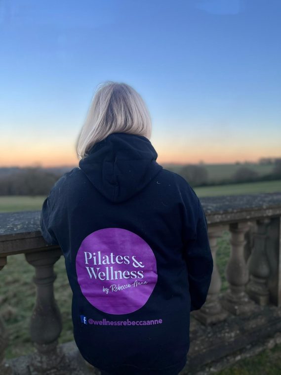 Rebecca is facing a relaxing sunset wearing a hoodie that has the Pilates & Wellness by Rebecca Anne logo on.